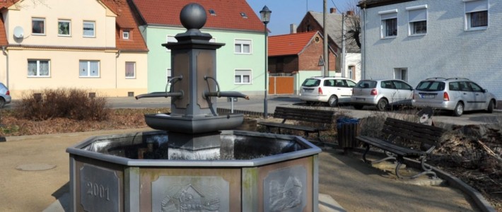 Der historische Brunnen von Löbnitz steht im Zentrum des Bildes. Um den Brunnen herum sind mehrere Bänke platziert, die zum Verweilen einladen. Im Hintergrund sieht man Häuser und einige parkende Autos um den ruhigen Dorfplatz.